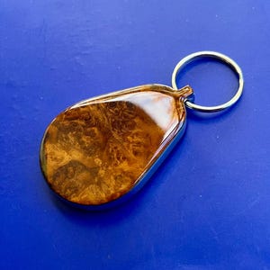 May include: A teardrop-shaped keychain featuring a polished brown burl wood inlay encased in a smooth, glossy resin finish. The wooden piece is framed by a silver-toned metal border and is attached to a circular silver-toned metal split ring.