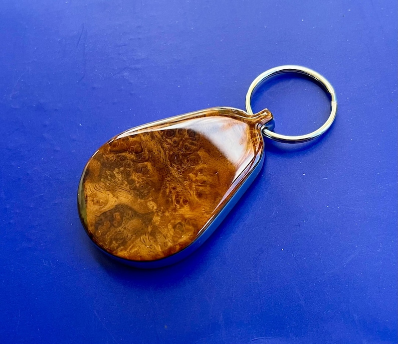 May include: A teardrop-shaped keychain featuring a polished brown burl wood inlay encased in a smooth, glossy resin finish. The wooden piece is framed by a silver-toned metal border and is attached to a circular silver-toned metal split ring.