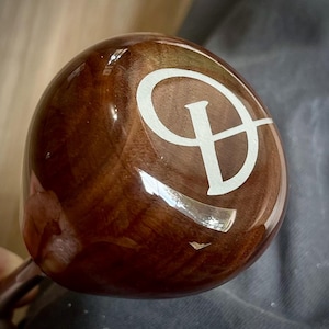 Puede incluir: Un objeto de madera redondeado y pulido con un acabado suave y brillante que presenta una letra D blanca prominente grabada o impresa en la superficie. La madera muestra un veteado marrón oscuro con remolinos naturales y reflejos.