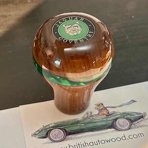 Pode incluir: Um manípulo de mudanças em madeira polida com acabamento brilhante, apresentando um emblema circular no topo com o texto Jaguar Coventry e um logótipo prateado de cabeça de jaguar. Uma faixa verde translúcida circunda o meio do manípulo.