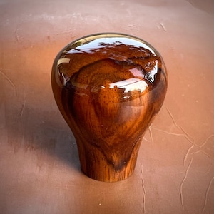 Puede incluir: Un pomo de palanca de cambios redondeado y pulido, fabricado en madera de color marrón oscuro con un patrón de veta visible y arremolinado. La superficie presenta un acabado brillante y reflectante que resalta la textura natural y los tonos ámbar.