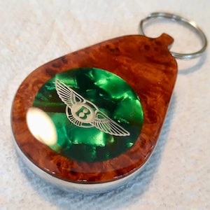 May include: A teardrop-shaped keychain featuring a polished brown burl wood frame surrounding a circular, vibrant green inlay. Centered on the green inlay is a silver-toned metallic emblem depicting a winged letter B logo with extended wings.