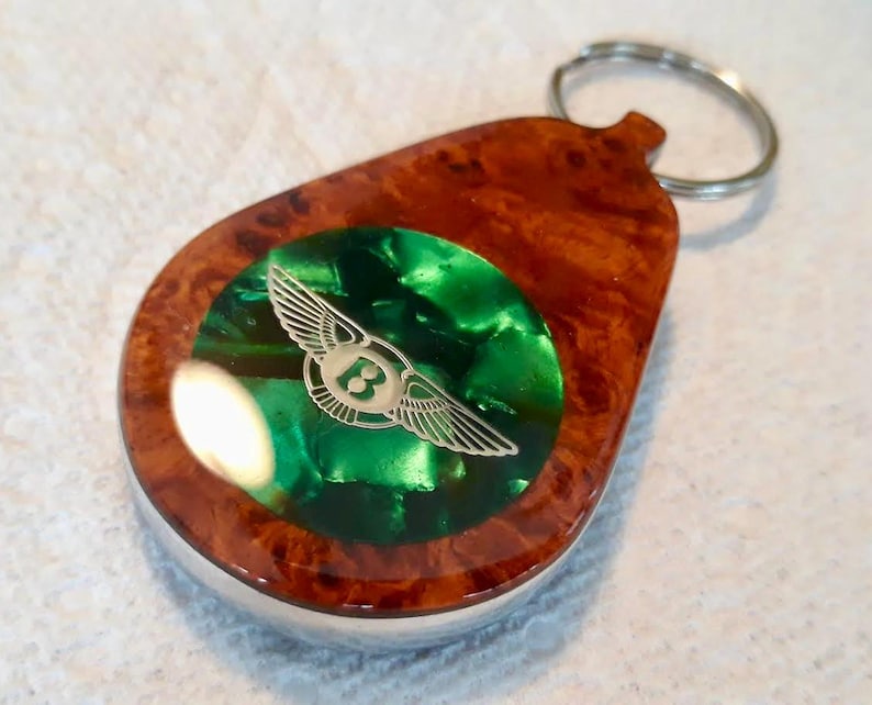 May include: A teardrop-shaped keychain featuring a polished brown burl wood frame surrounding a circular, vibrant green inlay. Centered on the green inlay is a silver-toned metallic emblem depicting a winged letter B logo with extended wings.