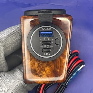 Puede incluir: Un cargador de coche con acabado de veta de madera. El cargador tiene un bisel negro con un puerto USB etiquetado QC3.0, dos puertos PD y un puerto DC. El cargador también tiene una cubierta abatible negra.