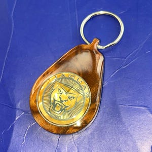May include: A teardrop-shaped keychain featuring a polished brown wood-grain finish and a circular gold-toned metal emblem in the center. The emblem displays the Jaguar brand logo, which includes the word JAGUAR and a roaring jaguar head.