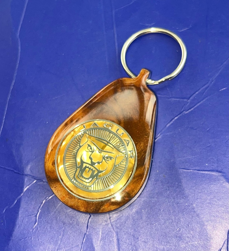 May include: A teardrop-shaped keychain featuring a polished brown wood-grain finish and a circular gold-toned metal emblem in the center. The emblem displays the Jaguar brand logo, which includes the word JAGUAR and a roaring jaguar head.