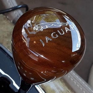 May include: A wooden Jaguar gear shift knob with a glossy finish. The Jaguar logo and text are etched in silver. The knob is a rich brown color and reflects light.