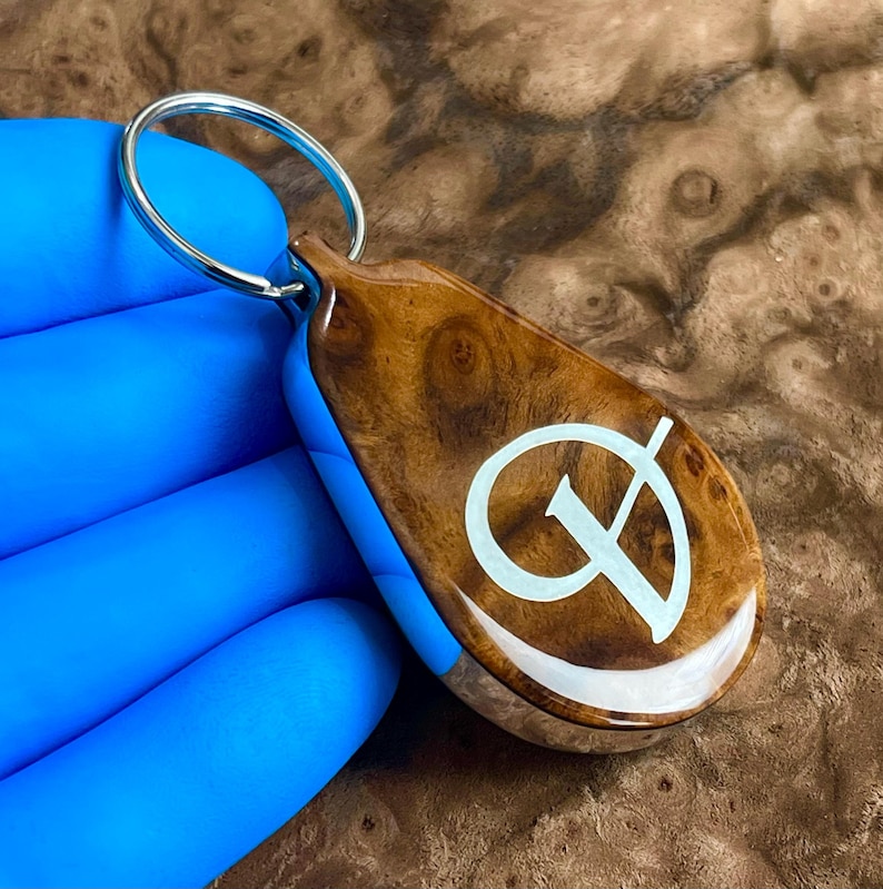May include: A teardrop-shaped keychain featuring a polished brown wood grain finish with a smooth, glossy surface. A white, stylized letter D is inlaid into the center of the wood. The keychain includes a circular silver metal ring for attachment.
