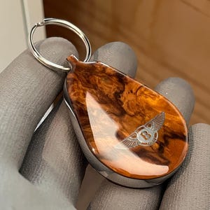 May include: A teardrop-shaped keychain featuring a polished, swirling brown wood grain finish with a glossy surface. The center displays a silver-toned engraved Bentley logo with wings and the letter B. A circular silver metal ring is attached at the top.