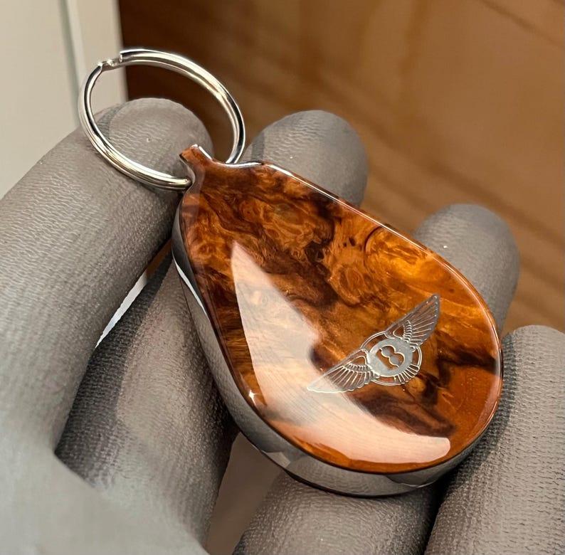 May include: A teardrop-shaped keychain featuring a polished, swirling brown wood grain finish with a glossy surface. The center displays a silver-toned engraved Bentley logo with wings and the letter B. A circular silver metal ring is attached at the top.