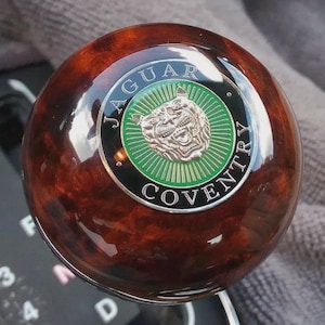May include: A wooden Jaguar gear stick knob with a polished finish. The knob has a silver emblem with a Jaguar head on a green background, encircled by the words "JAGUAR" and "COVENTRY" in a circular layout.