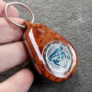 May include: A teardrop-shaped keychain featuring a polished burl wood finish and a central circular metal emblem. The emblem displays a stylized blue jaguar head logo with the text JAGUAR engraved in a silver border, attached to a silver metal ring.