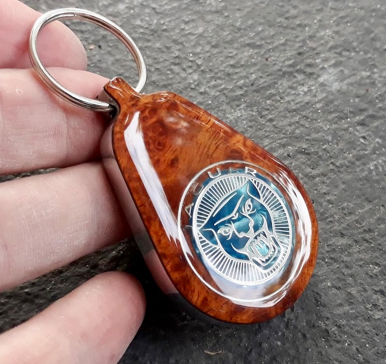 May include: A teardrop-shaped keychain featuring a polished burl wood finish and a central circular metal emblem. The emblem displays a stylized blue jaguar head logo with the text JAGUAR engraved in a silver border, attached to a silver metal ring.