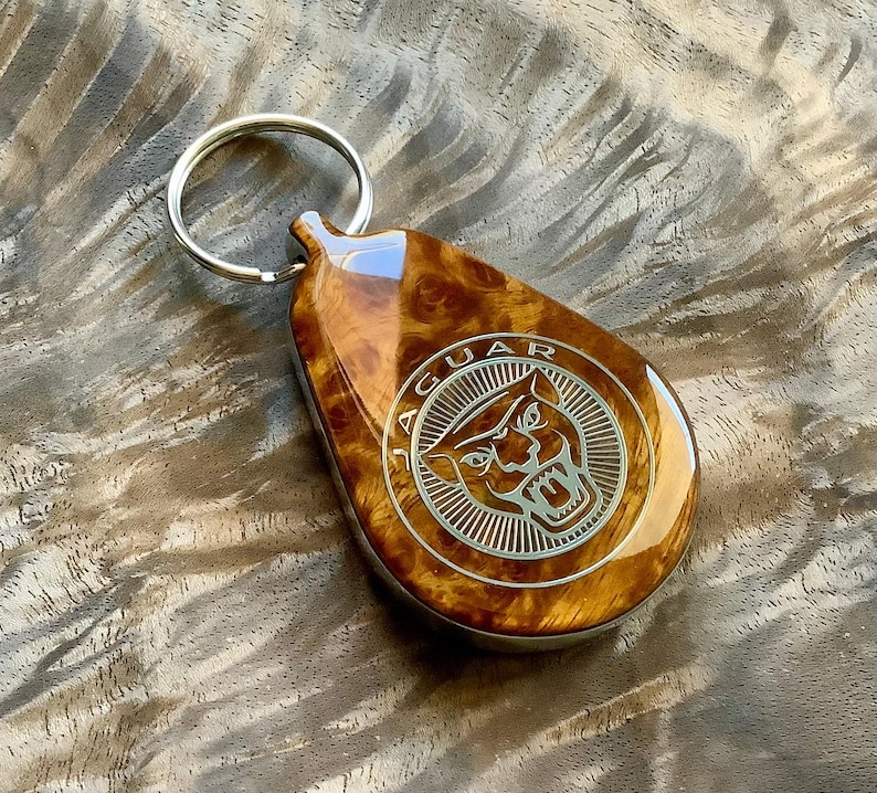 May include: A teardrop-shaped keychain featuring a polished, swirling brown burl wood finish and a silver-toned metal inlay. The inlay displays the Jaguar brand logo, which consists of a stylized roaring jaguar head inside a circular border.