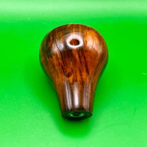 Op de afbeelding: Een gepolijste houten versnellingspookknop, donkerbruin met zichtbare houtnerf. De knop heeft een gat aan de bovenkant en een metalen basis. De achtergrond is groen.