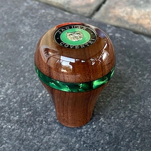 Pode incluir: Um manípulo de mudanças em madeira polida com um topo liso e arredondado, apresentando um emblema circular com um logótipo Jaguar verde e o texto JAGUAR COVENTRY. Uma faixa verde translúcida e vibrante circunda o centro da madeira castanha.