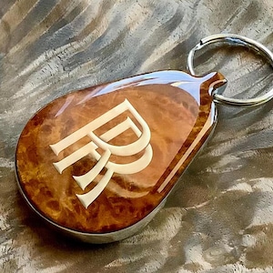 May include: A teardrop-shaped keychain featuring a polished, glossy brown burl wood inlay with a prominent, light-colored double R monogram embedded in the center. The keychain is attached to a circular silver-toned metal split ring.