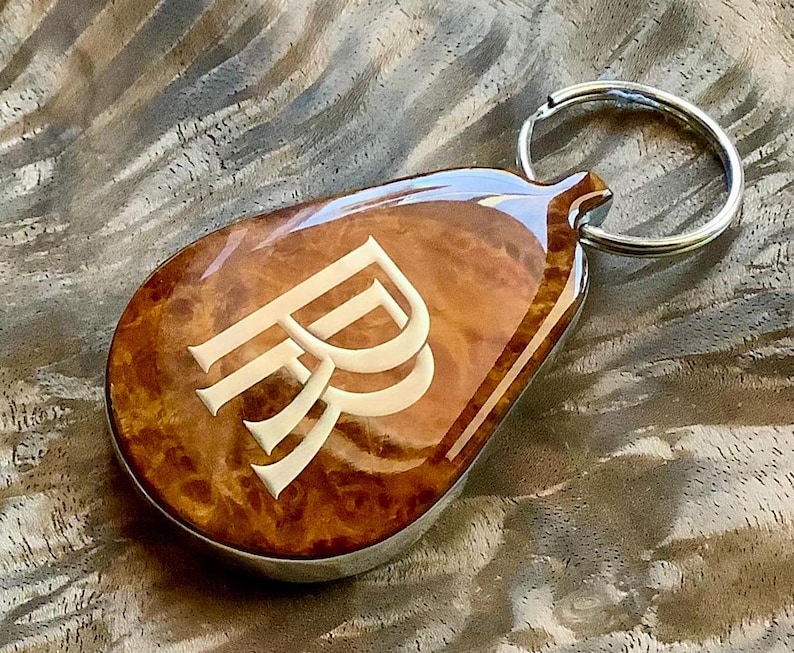 May include: A teardrop-shaped keychain featuring a polished, glossy brown burl wood inlay with a prominent, light-colored double R monogram embedded in the center. The keychain is attached to a circular silver-toned metal split ring.