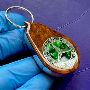 May include: A teardrop-shaped keyring with a Mercedes-Benz logo. The keyring has a silver ring, a wooden body, and a green and silver emblem. The wood has a natural grain pattern.