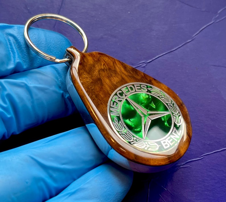 May include: A teardrop-shaped keychain featuring a polished wood grain body and a central circular emblem. The emblem displays the silver Mercedes-Benz three-pointed star logo set against a vibrant green background, surrounded by silver text.