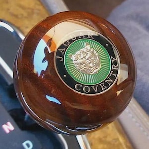 May include: A polished, round, dark brown wood gear shift knob featuring a circular emblem in the center. The emblem displays a silver jaguar head on a green background, surrounded by a black border with the text JAGUAR COVENTRY in silver.