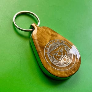 以下が含まれることがあります： 銀色の金属リングが付いた、涙型の木製キーホルダー。キーホルダーには、銀色の金属の円の中にジャガーのロゴが付いています。木材は、光沢のある仕上がりで、金色がかった茶色をしています。