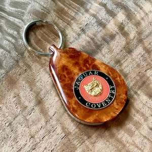 May include: A teardrop-shaped keychain featuring a polished burl wood finish and a circular emblem. The emblem displays a gold-colored jaguar head on a red background, surrounded by a black border with the text Jaguar Coventry in gold lettering.