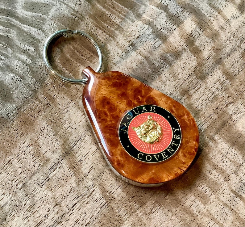 May include: A teardrop-shaped keychain featuring a polished burl wood finish and a circular emblem. The emblem displays a gold-colored jaguar head on a red background, surrounded by a black border with the text Jaguar Coventry in gold lettering.