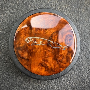 May include: Circular car emblem with a wood grain finish and a silver Jaguar logo. The emblem has a black border and a glossy, reflective surface. The wood grain features shades of brown and orange.