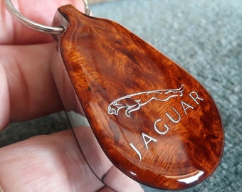 May include: A teardrop-shaped keychain featuring a polished brown wood grain inlay with a silver-toned metal border. The surface displays an engraved silver Jaguar logo and the word JAGUAR in capital letters, attached to a circular metal ring.