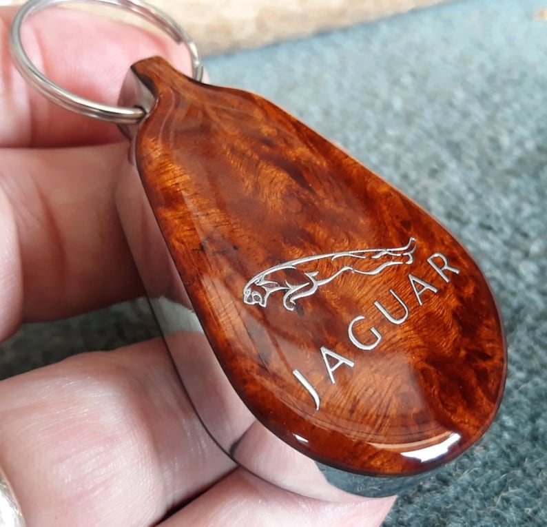 May include: A teardrop-shaped keychain featuring a polished, rich brown burl wood inlay set into a silver-toned metal frame. The wood surface is engraved with the silver Jaguar leaping cat logo and the word JAGUAR in capital letters.