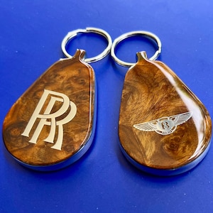May include: Two teardrop-shaped keychains with a polished wood grain finish and silver metal edges. One keychain features the double R logo, while the other displays a winged B emblem. Each includes a circular silver metal split ring for keys.