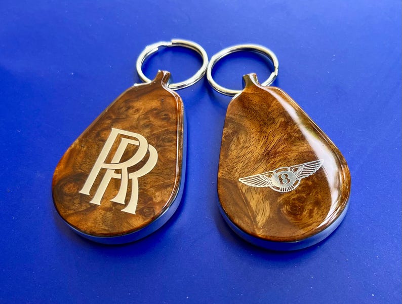 May include: Two teardrop-shaped keychains with a polished wood grain finish and silver metal edges. One keychain features the double R logo, while the other displays a winged B emblem. Each includes a circular silver metal split ring for keys.