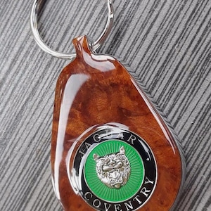 May include: A teardrop-shaped keychain featuring a polished brown burl wood finish and a circular emblem. The emblem displays a silver jaguar head on a green background, surrounded by a black border with the text JAGUAR COVENTRY in silver.