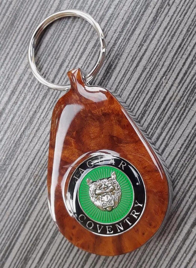May include: A teardrop-shaped keychain featuring a polished brown burl wood finish and a circular emblem. The emblem displays a silver jaguar head on a green background, surrounded by a black border with the text JAGUAR COVENTRY in silver.
