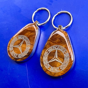 May include: Two teardrop-shaped keychains featuring a polished wood grain finish and a silver-toned metal border. Each keychain displays a laser-engraved Mercedes-Benz logo with the text Mercedes-Benz encircled by a laurel wreath design and a metal ring.