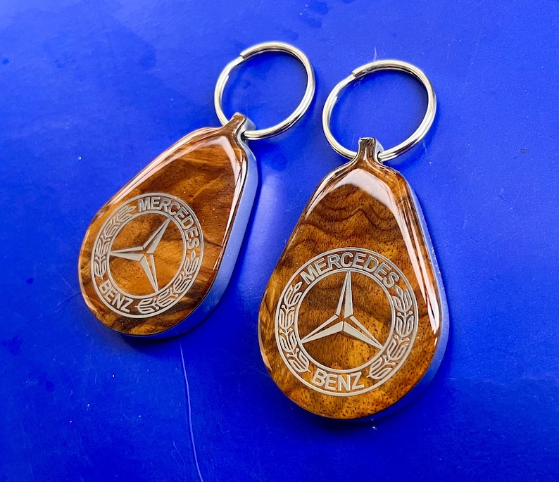 May include: Two teardrop-shaped keychains featuring a polished wood grain finish and a silver-toned metal border. Each keychain displays a laser-engraved Mercedes-Benz logo with the text Mercedes-Benz encircled by a laurel wreath design and a metal ring.