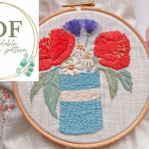 May include: Embroidered hoop art featuring a floral arrangement of red poppies, white daisies, and purple flowers in a blue and white striped vase. The embroidery is on a white linen fabric and is framed in a wooden hoop.