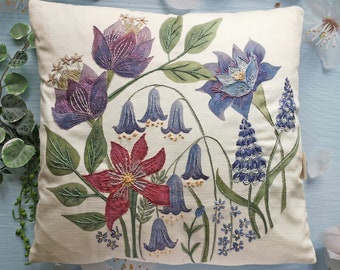 May include: A white cushion with embroidered floral designs. The cushion features a variety of flowers in shades of blue, purple, and red, including bellflowers, a large blue flower, and a red flower with a yellow centre.
