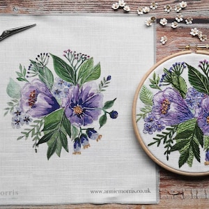 May include: A floral embroidery design featuring purple flowers and green leaves. The design is on a white fabric square and also displayed in a wooden embroidery hoop. A pair of scissors and the website address www.anniemorris.co.uk are also visible.