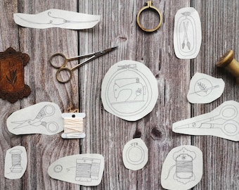 Op de afbeelding: Een assortiment naai- en borduurgereedschappen en -benodigdheden is gerangschikt op een houten oppervlak. De items omvatten scharen, borduurringen, klosjes garen en geborduurde ontwerpen. Een houten spoel en een decoratief frame zijn ook aanwezig.