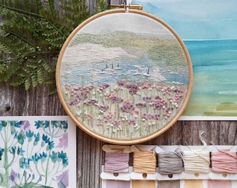 May include: A round embroidery hoop with a landscape scene featuring a body of water, sailboats, and a field of purple and pink flowers. Watercolor paintings and embroidery floss are also visible.