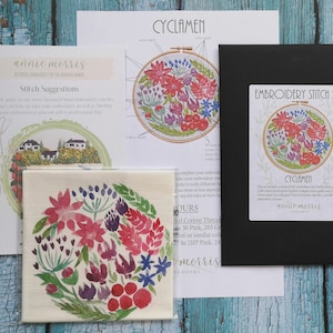 May include: A close-up of a embroidery kit for a floral design called "Cyclamen". The kit includes a printed pattern, embroidery floss, and a needle. The pattern features a circle of pink, red, and purple flowers with green leaves.
