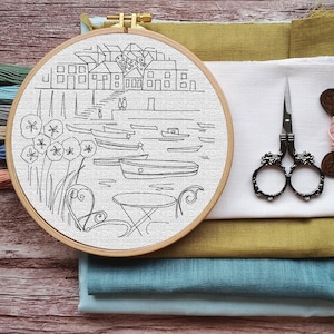 May include: A wooden embroidery hoop with a black and white line drawing of a waterfront scene. The scene includes buildings, boats, and a table with chairs. The hoop is on top of a stack of fabric in various shades of blue and green.