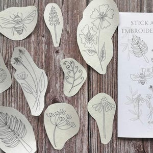May include: A selection of floral embroidery templates, featuring a butterfly, bee, and diverse flowers, displayed on a wooden background. A printed sheet with the text "Stick and Stitch Embroidery Templates" and "The Floral Set" is also visible.