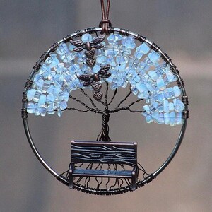 May include: A handcrafted, circular wire art piece featuring a tree design. The tree has a bench at its base and is adorned with light blue, gem-like beads. Two dragonfly charms are placed on the tree. The piece is suspended by a brown leather cord.