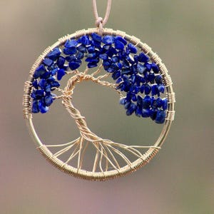 Puede incluir: Colgante artesanal con diseño de árbol, elaborado con alambre plateado. Las ramas del árbol están adornadas con piedras preciosas azules, engarzadas en un marco circular. El colgante cuelga de un cordón de color neutro.