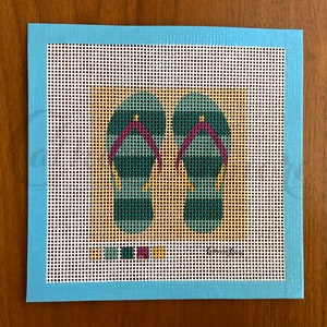 May include: A needlepoint canvas with a design of two teal and purple flip flops on a tan background. The canvas is surrounded by a light blue border.