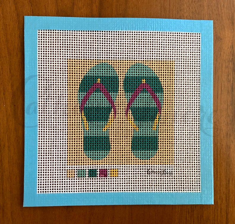May include: A needlepoint canvas with a design of two teal and purple flip flops on a tan background. The canvas is surrounded by a light blue border.