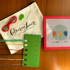 May include: A needlepoint kit with a red, yellow, and blue balloon design. The kit includes a canvas, embroidery floss, a needle, a threader, and scissors. The kit is packaged in a white canvas bag with the text "Catherine Senore Modern Needlepoint" printed on it.